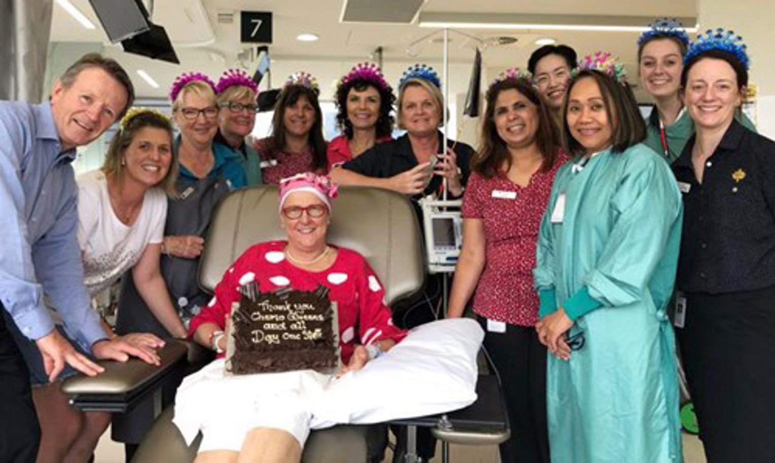 Maryanne sitting in chemo chair with nurses gathered around. She's holding a cake that says 'thank you chemo queens and all day one staff'
