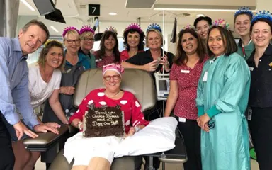Maryanne sitting in chemo chair surrounded by nurses. she's holding a cake that says 'thank you chemo queens and all day one staff'