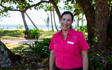 Jessie Chenery is standing in a sunny park with the ocean behind her. She is wearing a bright, pink shirt and smiling at the camera.
