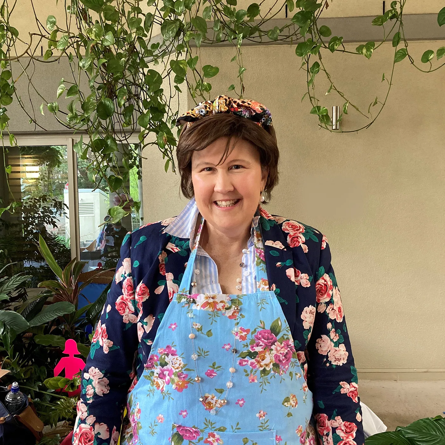 Ros has short, brown hair and is wearing a floral apron. She is smiling and looking straight at the camera.