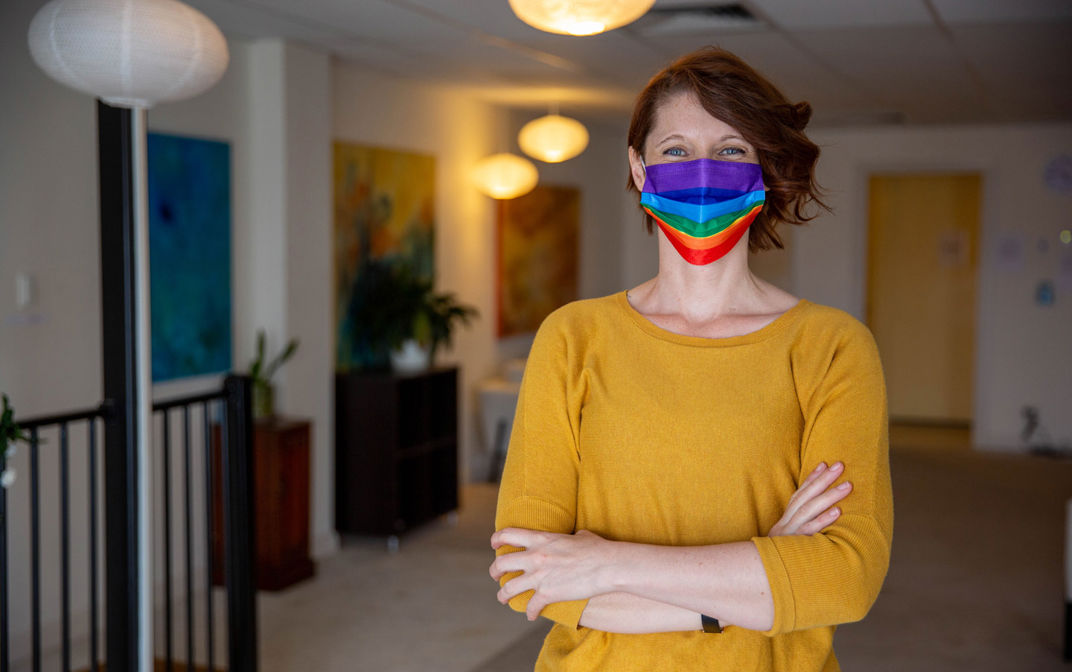 A woman is wearing a yellow jumper and stands with her arms crossed. She is wearing a rainbow face mask and looking at the camera.
