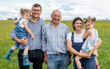 A family of multiple generations including young children are standing in a paddock on a farm looking at the camera
