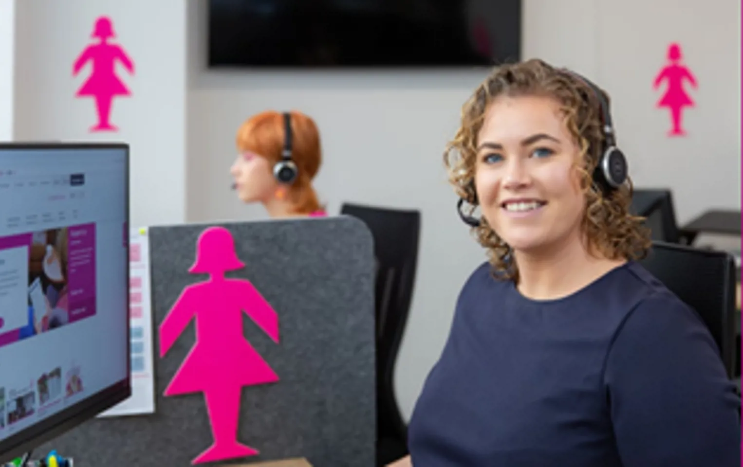 A young woman with brown curly hair is smiling at the camera from her office cubicle. She is wearing headphones and in the background are several Pink Lady cutouts  