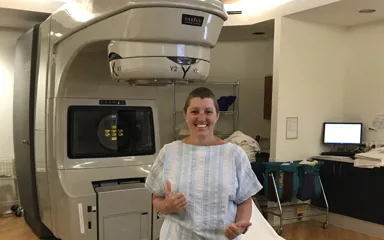 Megan stands beside hospital scanning machine. She is smiling and giving two thumbs up