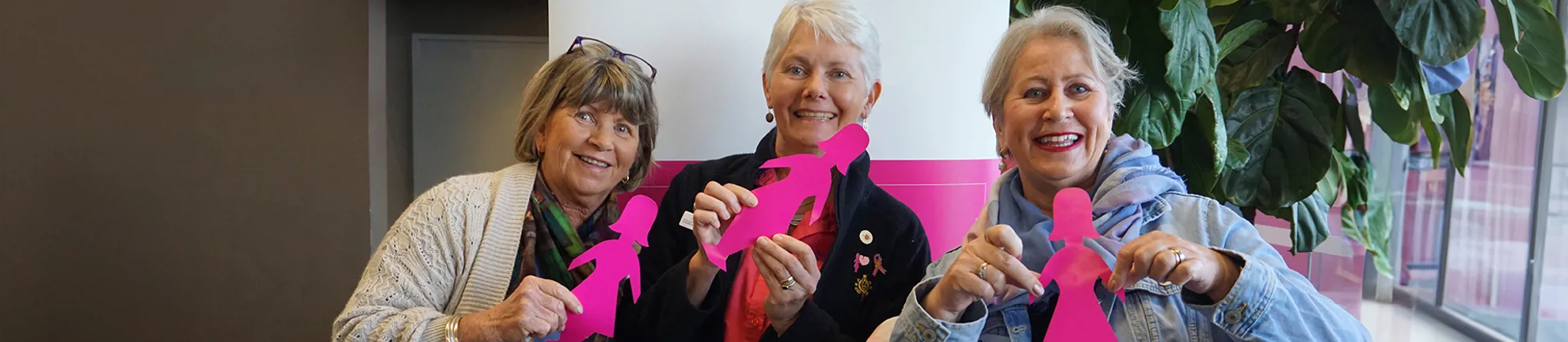 Three women are sitting next to each other holding BCNA pink lady silhouettes and smiling at the camera
