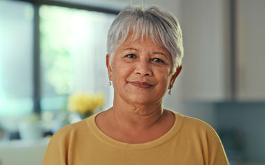 A senior woman at home looking at the camera. She has grey hair, olive skin and is wearing a yellow top.