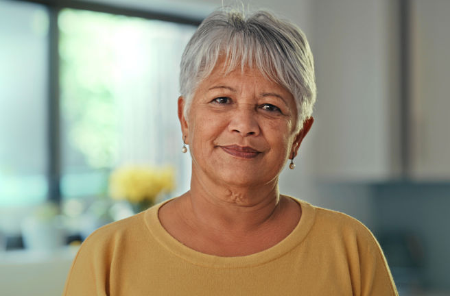 A lady is standing in a light-filled room. She has short, grey hair and a looking straight at the camera. She is smiling.