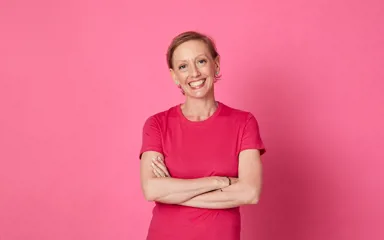 Kath smiling at the camera with her arms crossed. She's wearing a pink top and standing in front of a pink backdrop