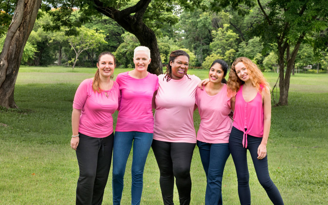 Five women all dressed in pink tops are standing in a park with their arms around each other