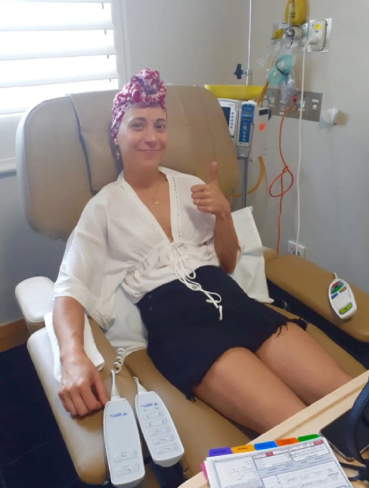 Lauren sitting in hospital chair receiving treatment, there are monitors behind her, she wears a pink and white headscarf and is smiling at the camera with her thumbs up