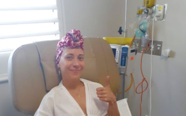 Lauren sitting in a hospital chair receiving treatment. She's wearing a pink and white headscarf and smiling with her thumbs up