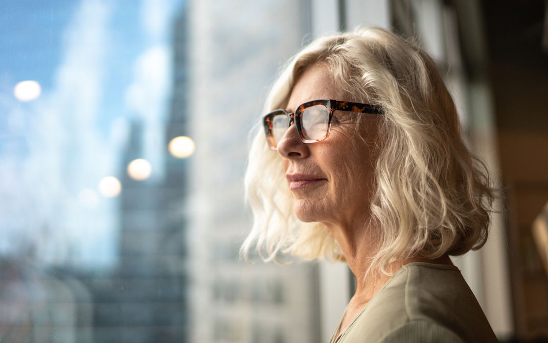 A mature woman is standing by a window looking out at the view of a city deep in thought