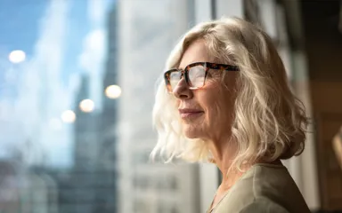 A mature woman is standing by a window looking out at the view of a city deep in thought