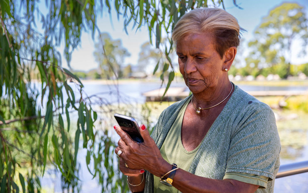A woman, wearing a light blue t-shirt, is standing next to a tree with a large river behind her. She is looking at a mobile phone in her hands.