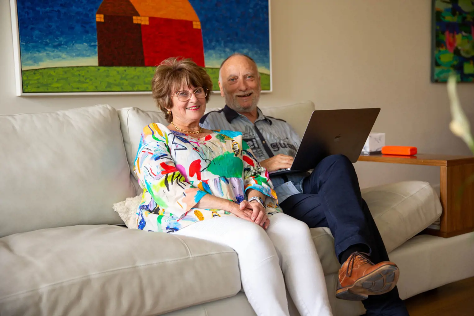 Photo of BCNA Member Harry, who has a white beard, with his wife Yvonne, who has auburn hair and is wearing a multicoloured shirt. They are sitting on a cream sofa. Harry has a laptop on his knees, and they are both smiling