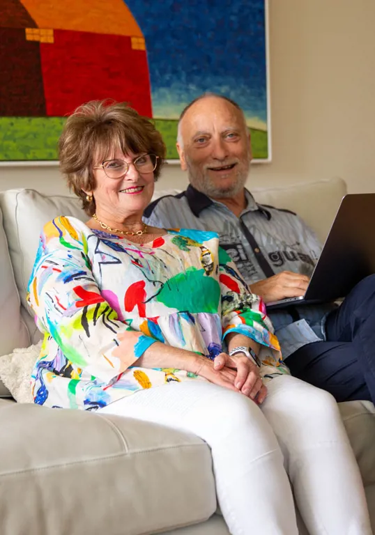 Photo shows an older man and woman sitting together on a cream-coloured couch, smiling. The man has a laptop open on his lap