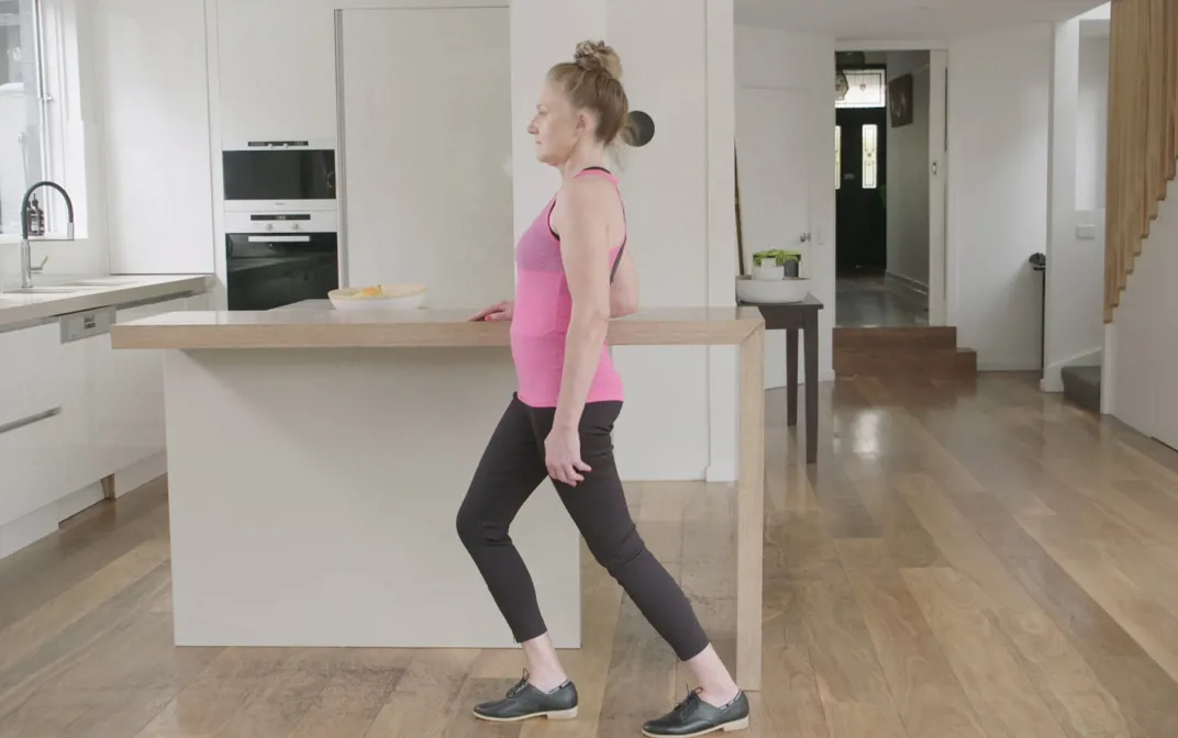 A woman is exercising in her kitchen, standing against her kitchen bench