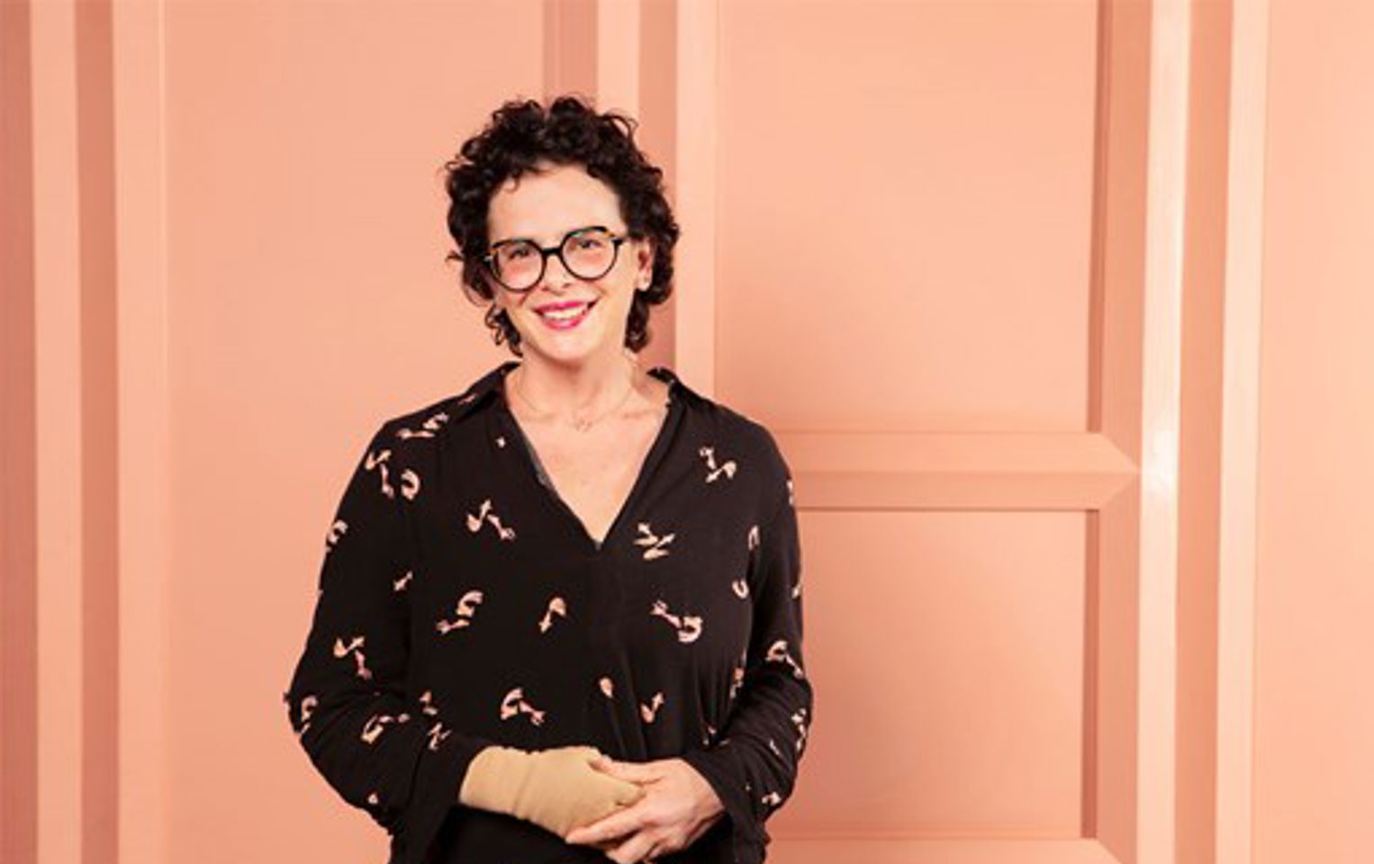 Photo of a smiling woman (Dr Na'ama Carlin) who is wearing glasses and pink lipstick. Her hair is short, dark and curly and she is wearing a dark dress, against a light pink background