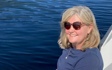 A woman with blonde hair, wearing blue jeans, a blue t-shirt and sunglasses is sitting on the edge of a boat with her feeling dangling over a blue stretch of water. She is smiling at the camera